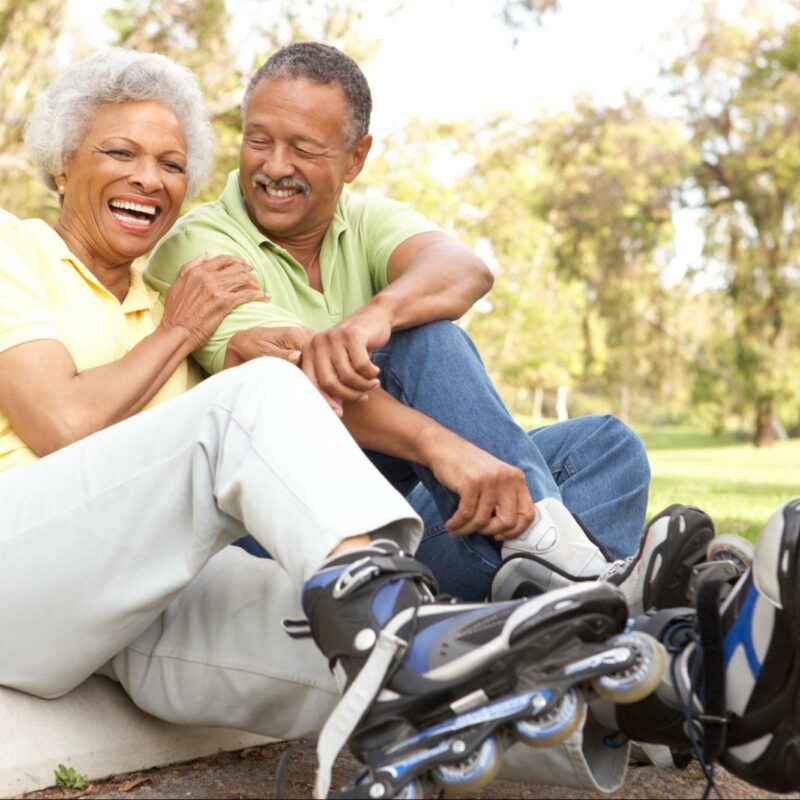 Two elderly people sitting and laughing together on a curb in a park, with inline skates on their feet, suggesting they have been active and are now resting.